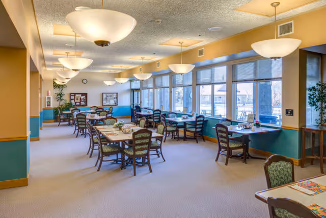 Bright assisted living dining room with multiple tables and chairs, large windows, and overhead pendant lights.