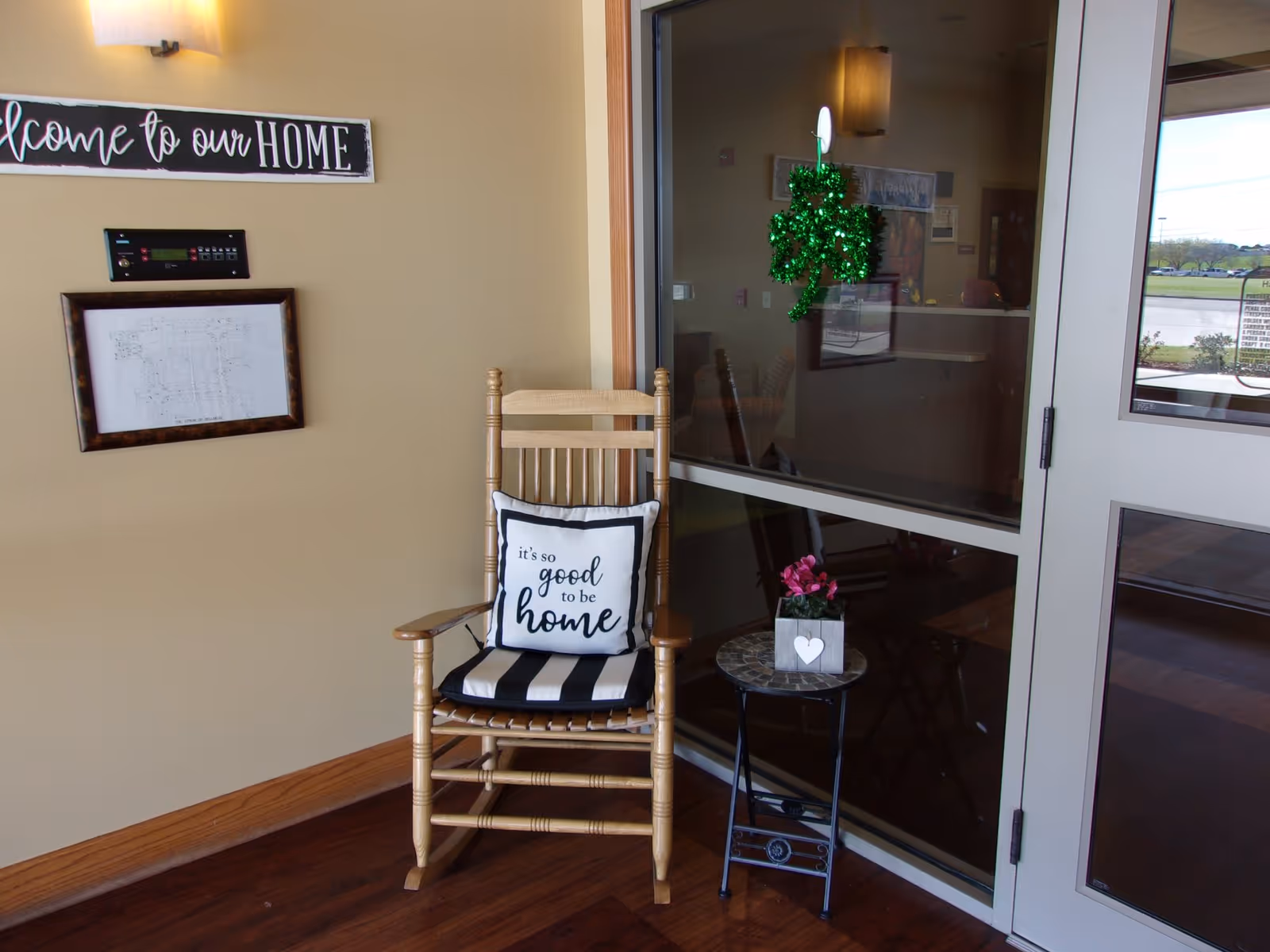 A cozy corner with a wooden rocking chair that has a black and white striped cushion and a pillow with the text 'it's so good to be home'. Next to the chair is a small round table with a potted pink flower. On the wall above the chair is a sign that says 'Welcome to our HOME' and a framed document. There is a glass door and window reflecting the interior and showing a green shamrock decoration hanging on the glass.