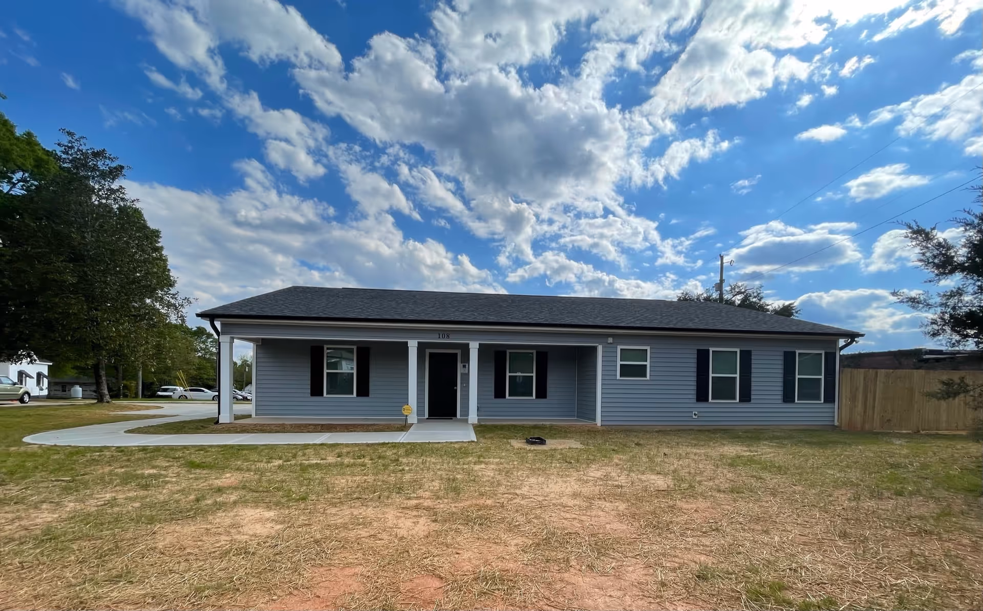Single-story residential building with gray siding, black shutters, and a black front door under a covered porch. The building is set against a partly cloudy blue sky with a grassy yard in the foreground.