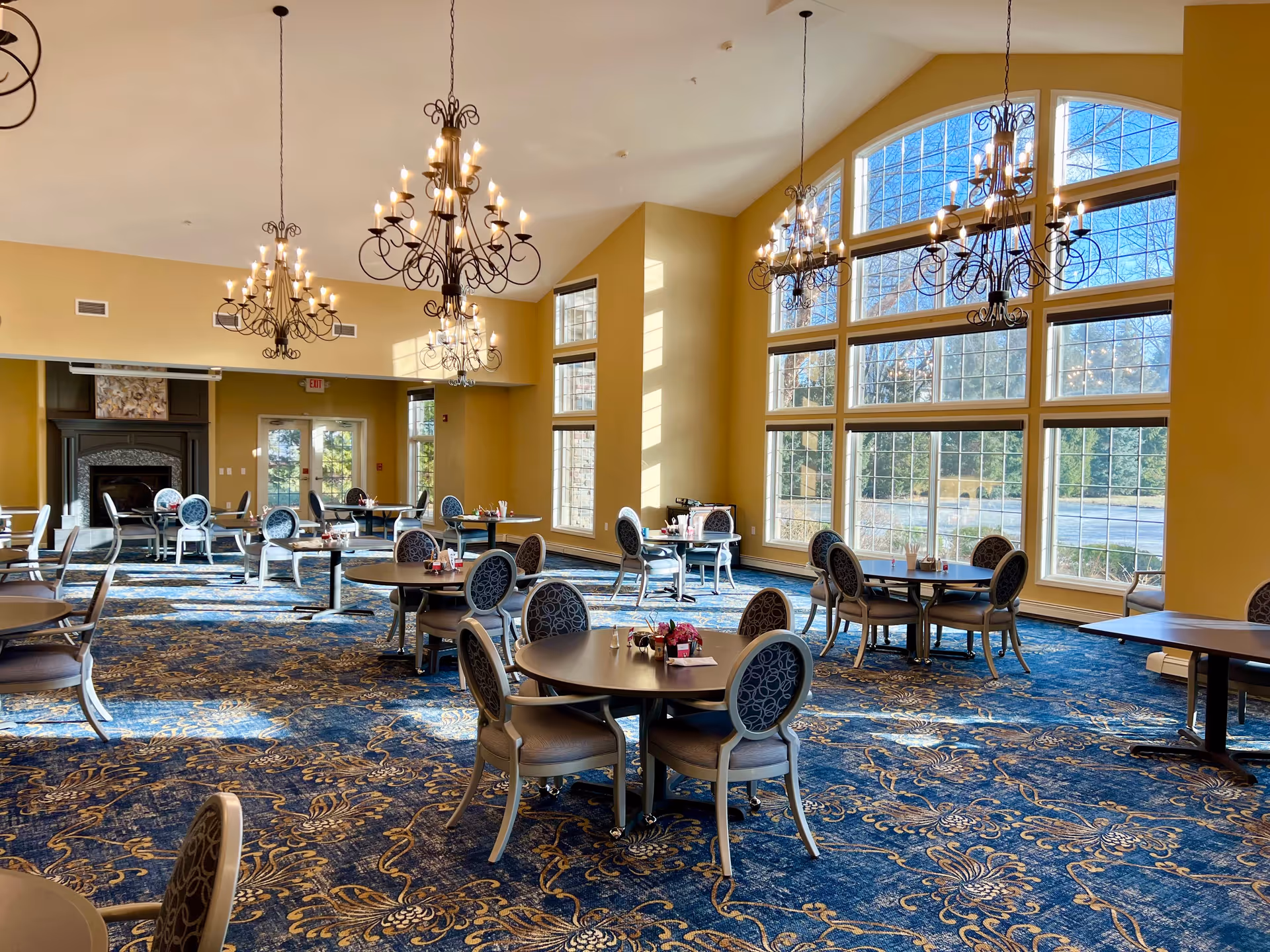 A spacious dining room with multiple round tables and chairs arranged neatly. The room features large arched windows allowing natural light to fill the space, ornate chandeliers hanging from the ceiling, and a patterned blue carpet with gold designs. There is a fireplace at the far end of the room.