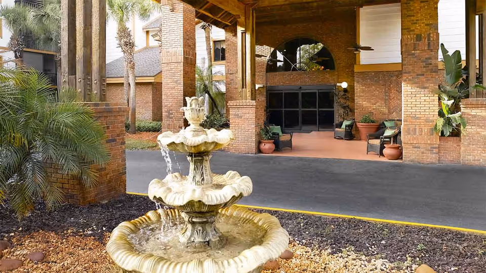 Outdoor view of the entrance to a senior living facility with a three-tiered water fountain in the foreground, surrounded by landscaping with plants and mulch. The building features brick pillars and a covered entryway with seating and potted plants.