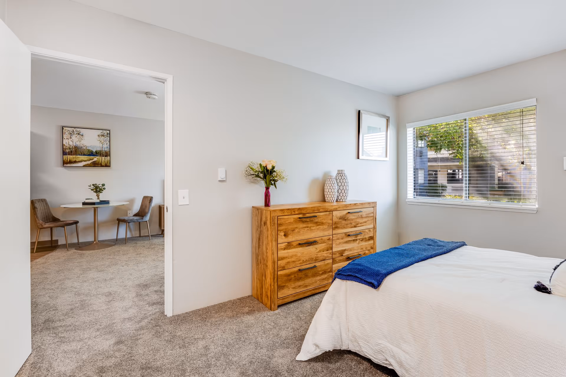 A bright bedroom with a wooden dresser topped with decorative vases and flowers, a bed with white bedding and a blue throw, and a window with blinds showing greenery outside. Through an open door, a small dining area with a round table, two chairs, and a framed landscape painting on the wall is visible.