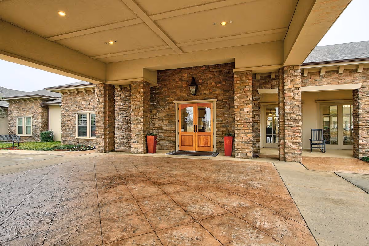 Covered entrance area of a building with stone pillars and walls, wooden double doors in the center flanked by two tall red planters, and a bench and rocking chair visible on the sides.