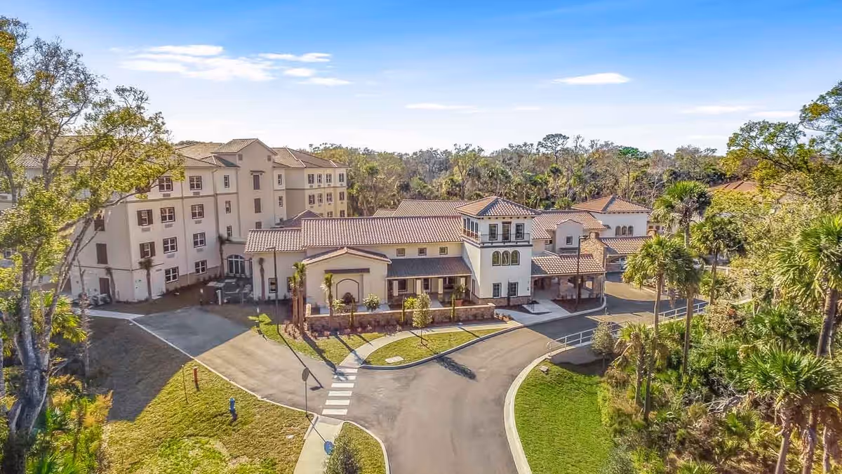 Aerial view of Tuscan Gardens of Palm Coast Senior Living facility showing a large multi-story building with a tiled roof surrounded by trees and greenery under a blue sky.