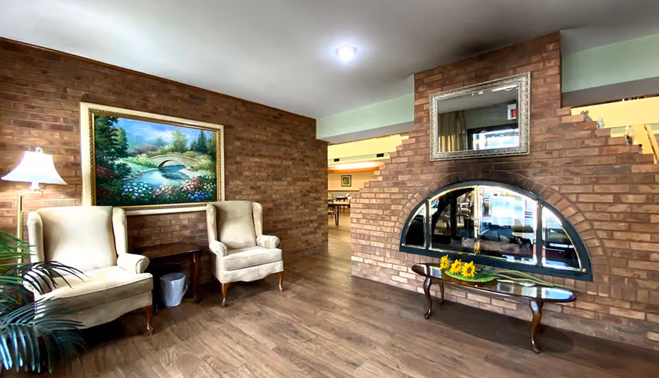 A cozy sitting area with two beige armchairs and a small wooden side table between them, placed against a brick wall with a framed painting of a garden and bridge. To the right, there is a large brick fireplace with a mirror above it and a wooden coffee table in front holding a decorative arrangement of sunflowers. The floor is wooden, and the space is warmly lit.