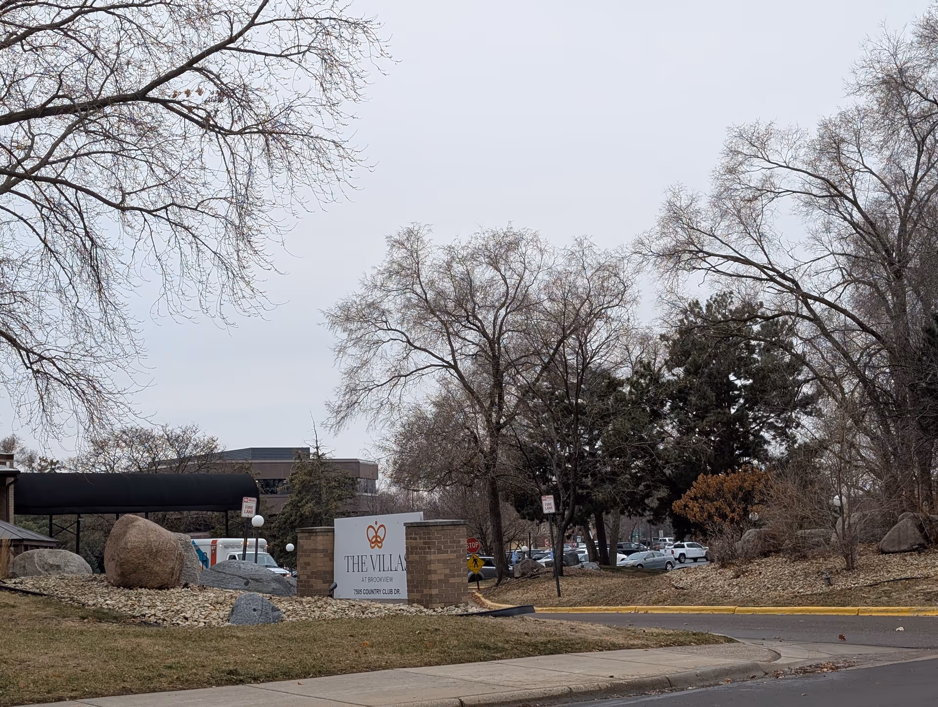 Outdoor view of the entrance to The Villas at Brookview senior living facility, showing a sign with the facility's name and address surrounded by rocks and trees with bare branches on a cloudy day.