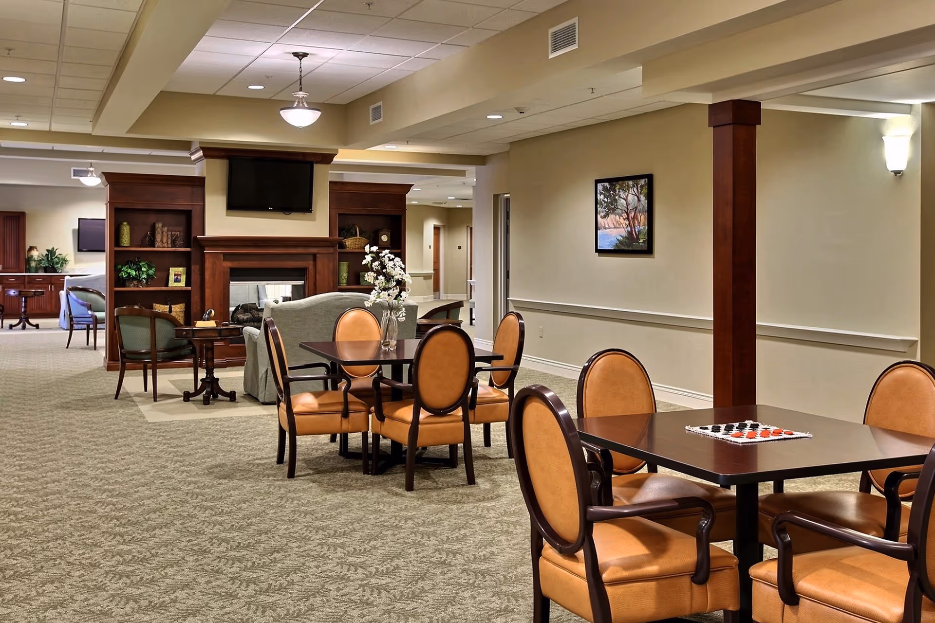 A spacious and well-lit common area in an assisted living facility featuring multiple tables with chairs, a fireplace with a mounted TV above it, and comfortable seating including armchairs and a sofa. The room has beige walls, carpeted floors, and decorative elements such as a framed painting and a vase with flowers on one of the tables.