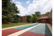 Shuffleboard courts in a courtyard beside a brick senior living building with trees and a partly cloudy sky.