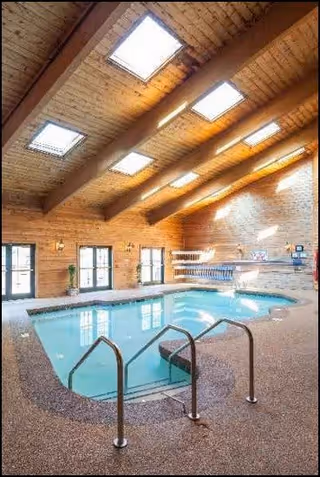 Indoor swimming pool area with a textured floor, metal handrails, and several skylights in a wooden ceiling. The walls are made of wood paneling with windows along one side, allowing natural light to enter the space.