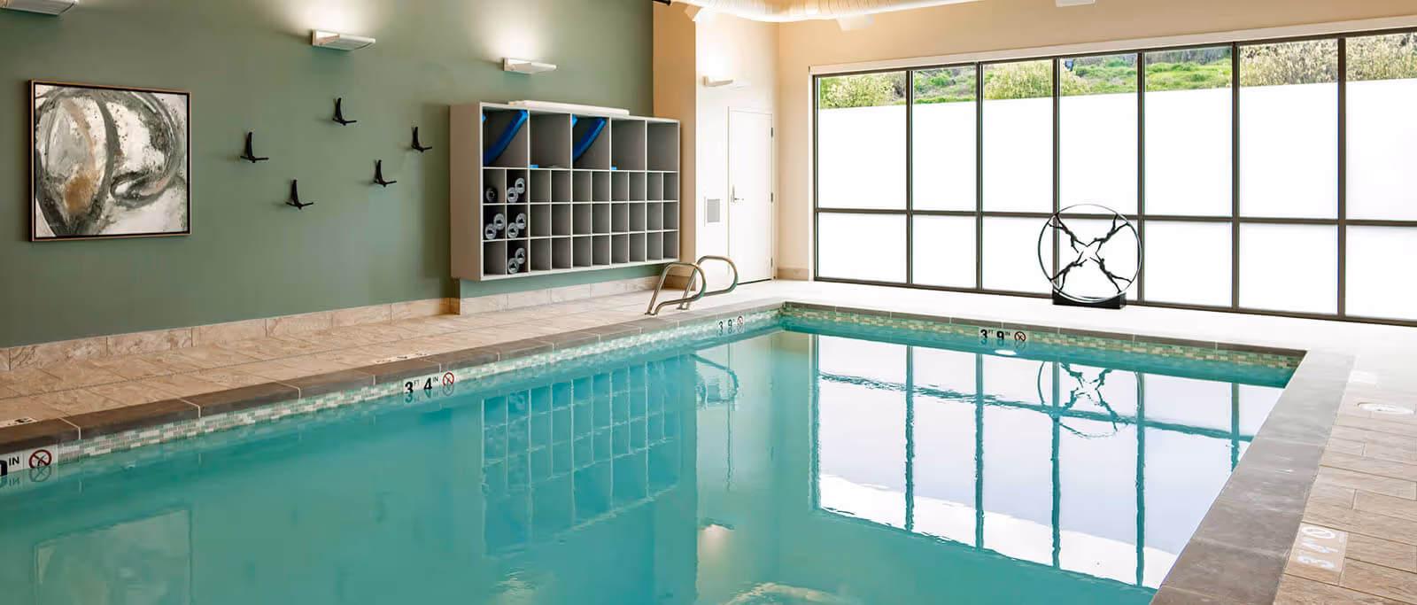 Indoor swimming pool with clear blue water, surrounded by tiled flooring and a green wall with hooks and cubby shelves holding rolled towels and pool equipment. Large windows let in natural light and offer a view of greenery outside.