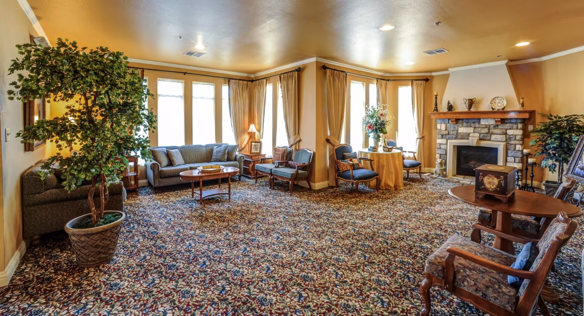 A spacious living room with patterned carpet, multiple seating areas including sofas and armchairs, a stone fireplace with decorative items on the mantel, large windows with beige curtains, and potted plants adding greenery to the room.