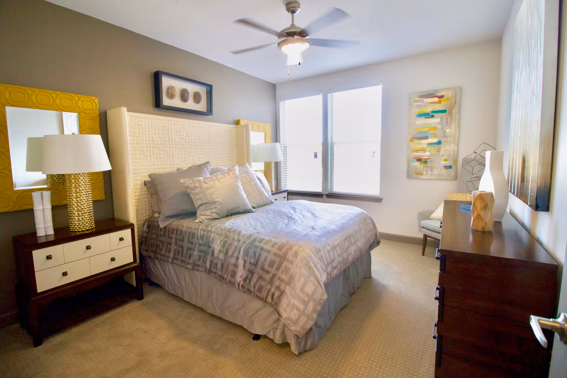 A well-lit bedroom with a large bed featuring a white padded headboard and gray patterned bedding. There are two yellow-framed mirrors on the wall behind the bed, each with a white lamp on a dark wooden nightstand. A ceiling fan with a light is mounted on the ceiling. The room has two large windows letting in natural light, a dark wooden dresser with decorative items, and a modern abstract painting on the wall.