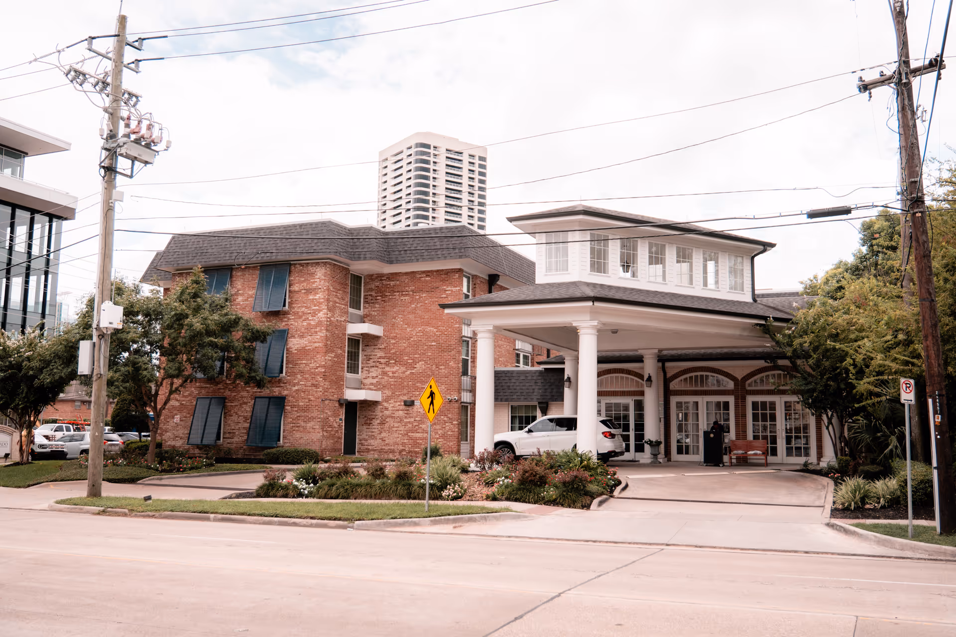 Exterior view of Silverado Hermann Park Memory Care Community showing a brick building with multiple windows and a covered entrance supported by white columns. A white SUV is parked under the entrance canopy. There are trees, shrubs, and a pedestrian crossing sign near the driveway.