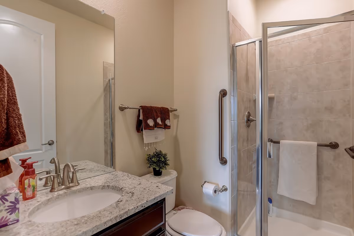 A clean and modern bathroom featuring a granite countertop with a sink, a large mirror, a toilet with a small potted plant on top, a towel rack with brown and white towels, and a glass-enclosed shower with beige tiles and a white towel hanging on the grab bar.