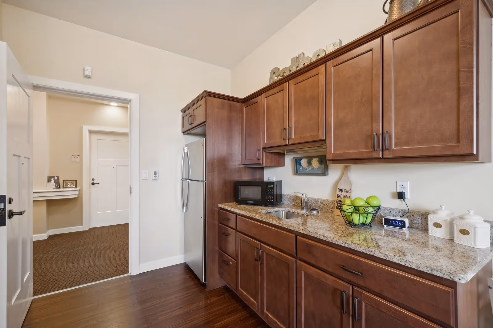 A kitchen area with wooden cabinets, granite countertops, a stainless steel refrigerator, a microwave, a small sink, and various decorative items including a bowl of green apples and a digital clock showing 11:35. The kitchen opens into a hallway with carpeted flooring and white doors.