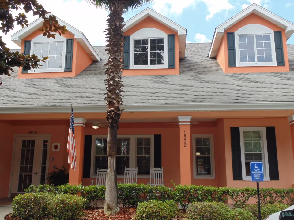 Front exterior view of a peach-colored assisted living facility building with three dormer windows on the roof, white rocking chairs on the porch, an American flag, and a handicapped parking sign in front.