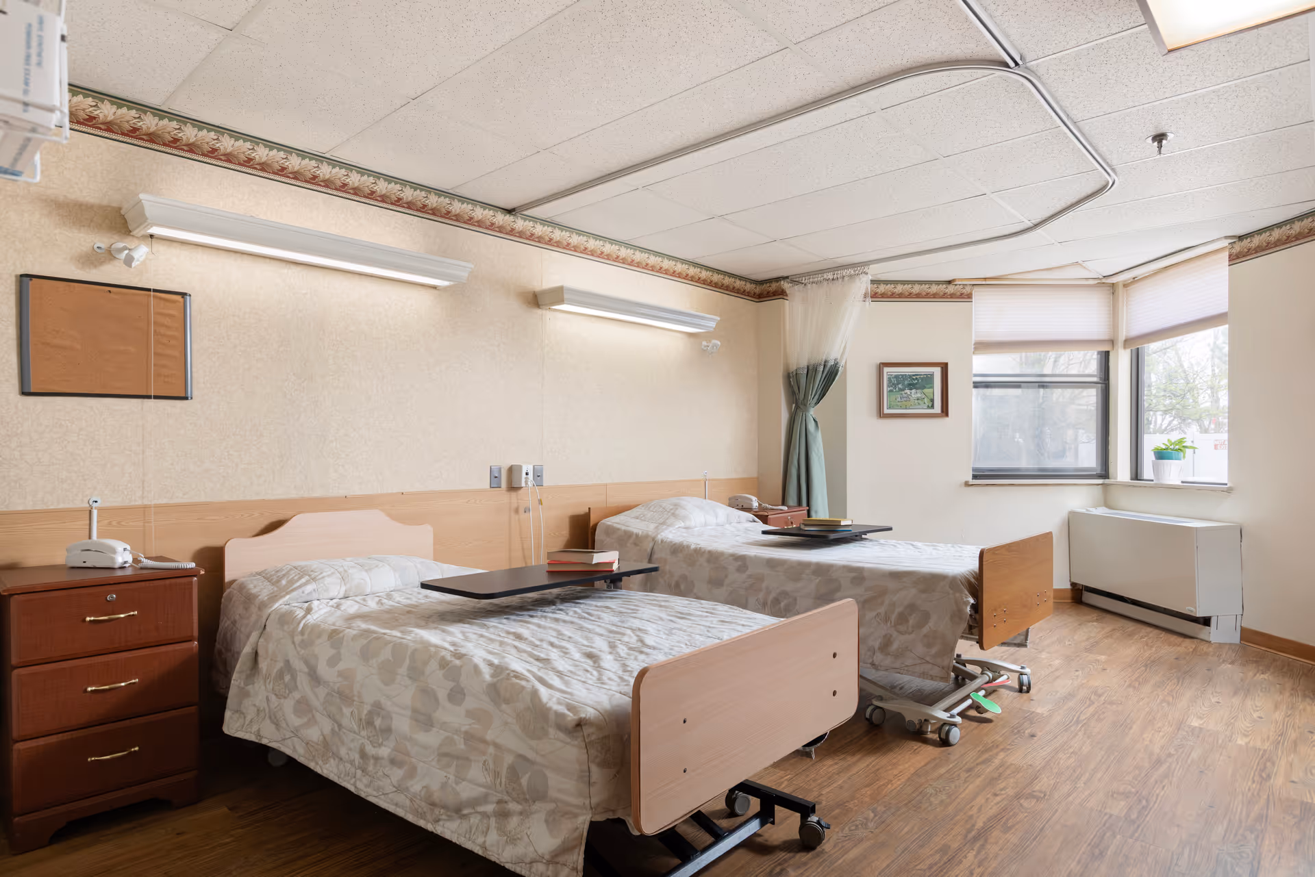 A bright room with two hospital-style beds covered in beige patterned bedspreads. Each bed has an overbed table with books on it. There is a wooden nightstand with a telephone on it between the beds. The room has wood flooring, beige walls with a floral border near the ceiling, and a large window with blinds and a small potted plant on the windowsill. A curtain is partially drawn near the corner of the room.