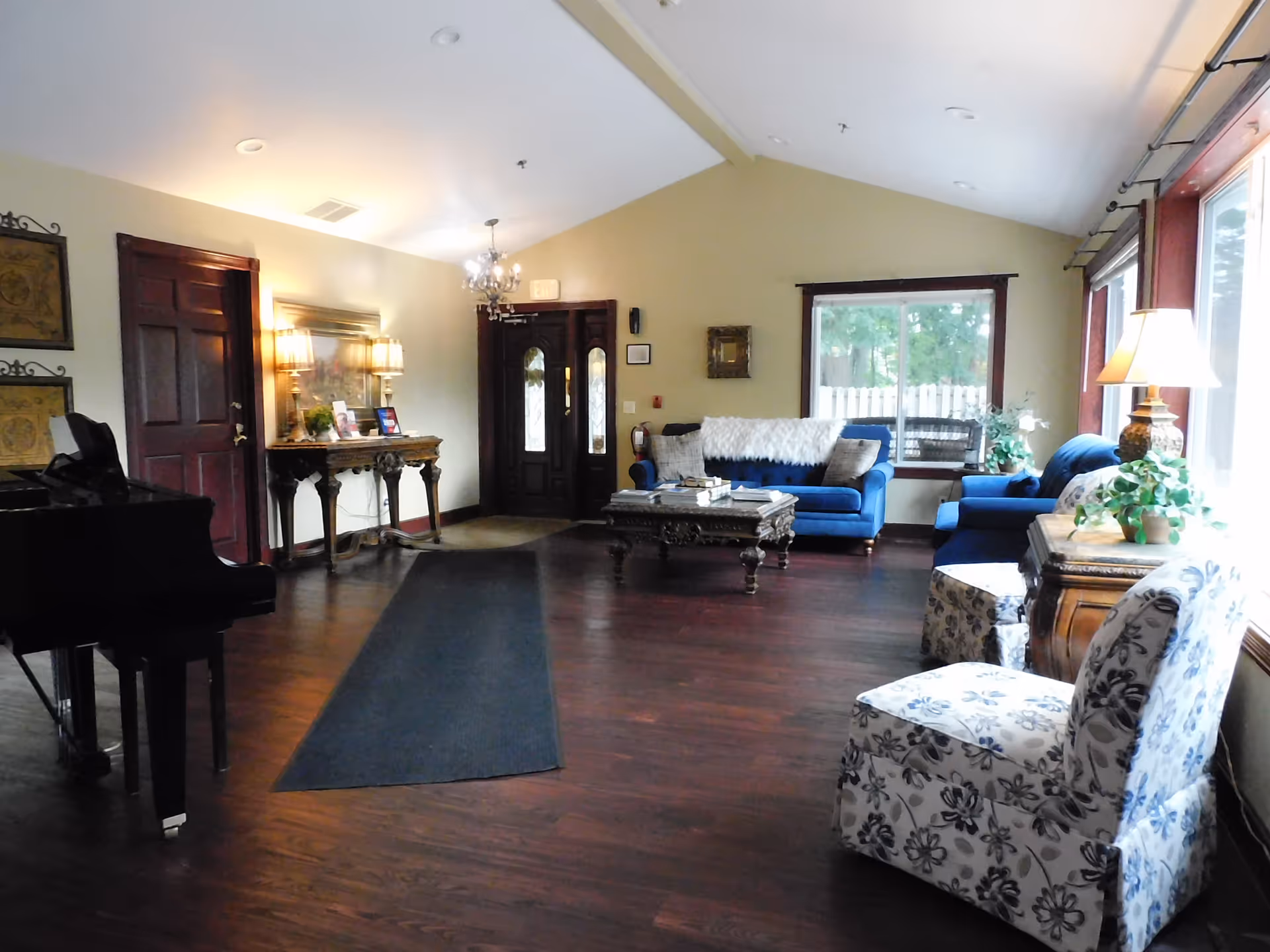 A spacious living room with dark wooden floors, a grand piano on the left, a decorative console table with lamps and framed pictures against the wall, and a seating area with blue sofas, a wooden coffee table, and floral-patterned chairs near large windows letting in natural light.