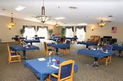 A spacious dining room with multiple square tables covered with blue tablecloths, each surrounded by wooden chairs with blue cushions. The room has large windows with curtains allowing natural light, ceiling lights, and some plants and decorations near the windows. An American flag is visible on the wall in the background.