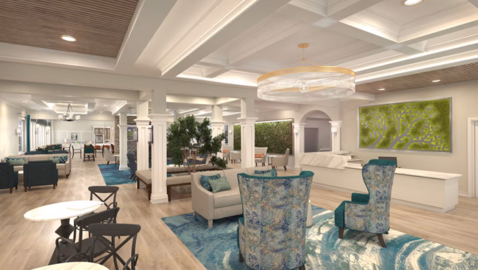 A spacious and well-lit senior living facility common area with comfortable seating including armchairs and sofas arranged on blue patterned rugs. The room features white columns, a modern chandelier, wood flooring, and green artwork on the walls. There are tables and chairs in the background, and some indoor plants adding a touch of nature.