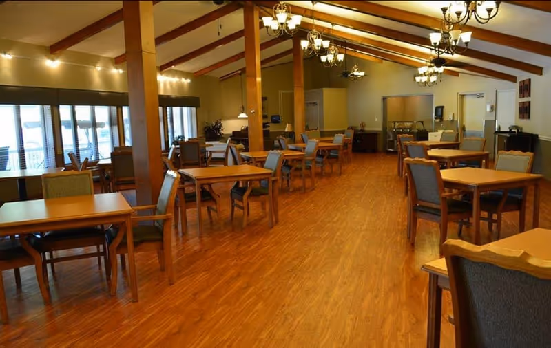 Interior view of a spacious dining room with wooden floors, multiple wooden tables and chairs arranged neatly, large windows with blinds on the left side, exposed wooden beams on the ceiling, and several chandeliers providing warm lighting.