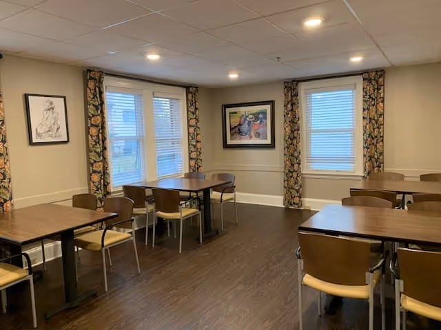 A dining room with several wooden tables and chairs arranged neatly. The room has three windows with floral patterned curtains, beige walls, and framed artwork hanging on the walls. The floor is dark wood, and the ceiling has recessed lighting.