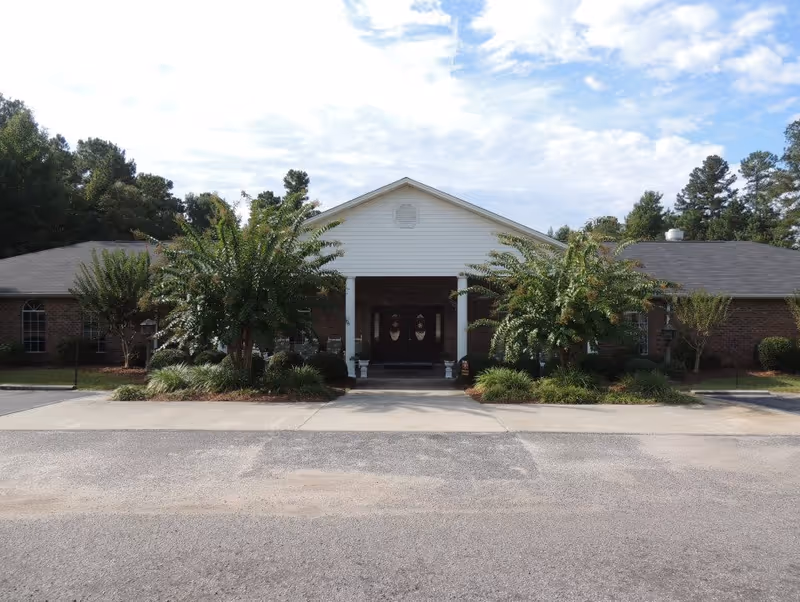 Front exterior view of a single-story brick building with a white gabled entrance supported by two white columns. There are two large bushes and landscaped greenery in front of the entrance, and the building is surrounded by trees under a partly cloudy sky.