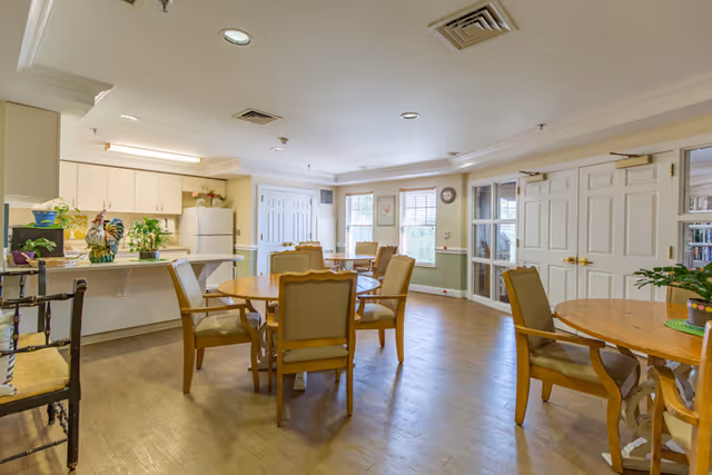 A bright and spacious dining area in a senior living facility featuring round wooden tables with cushioned chairs. The room has large windows allowing natural light to fill the space, light-colored walls with wainscoting, and a kitchen area with white cabinets and a refrigerator in the background. There are plants and decorative items on the counter and tables, and double doors leading to another room.