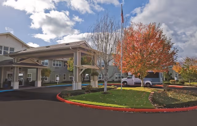 Exterior view of a senior living facility with a covered entrance, landscaped greenery, a tree with autumn-colored leaves, an American flag on a flagpole, and a white truck parked near the entrance under a partly cloudy sky.