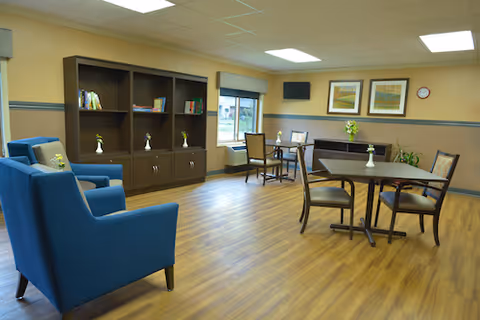 A bright communal lounge with blue armchairs, round tables and chairs, bookshelves, and framed art on the walls.