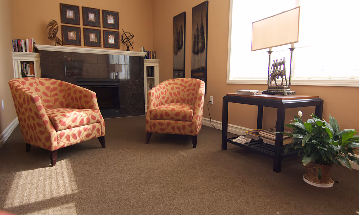 A cozy sitting area with two patterned armchairs facing each other on a brown carpet. Behind the chairs is a fireplace with a dark stone surround, flanked by built-in white shelves holding books and decorative items. On the right side, there is a wooden side table with a lamp and magazines underneath, next to a green potted plant. The room has beige walls and a large window letting in natural light.