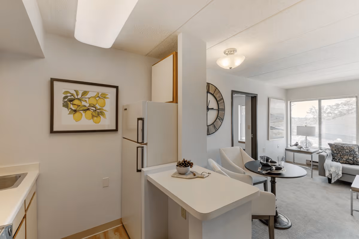 Interior view of a senior living facility showing a small kitchen area with a refrigerator, a framed picture of lemons on the wall, and a countertop with a bowl of grapes. Adjacent to the kitchen is a dining area with a round table set for two and two white chairs. In the background, there is a living room with a sofa, a side table with a lamp, a large window with blinds, and a large wall clock near a doorway.