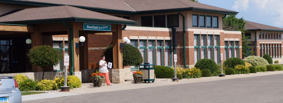 Exterior view of Eventide Heartland Care Center showing a brick building with large windows, landscaped bushes and flowers, a covered entrance with a sign reading 'Heartland Care Center', and a person standing near the entrance holding papers.