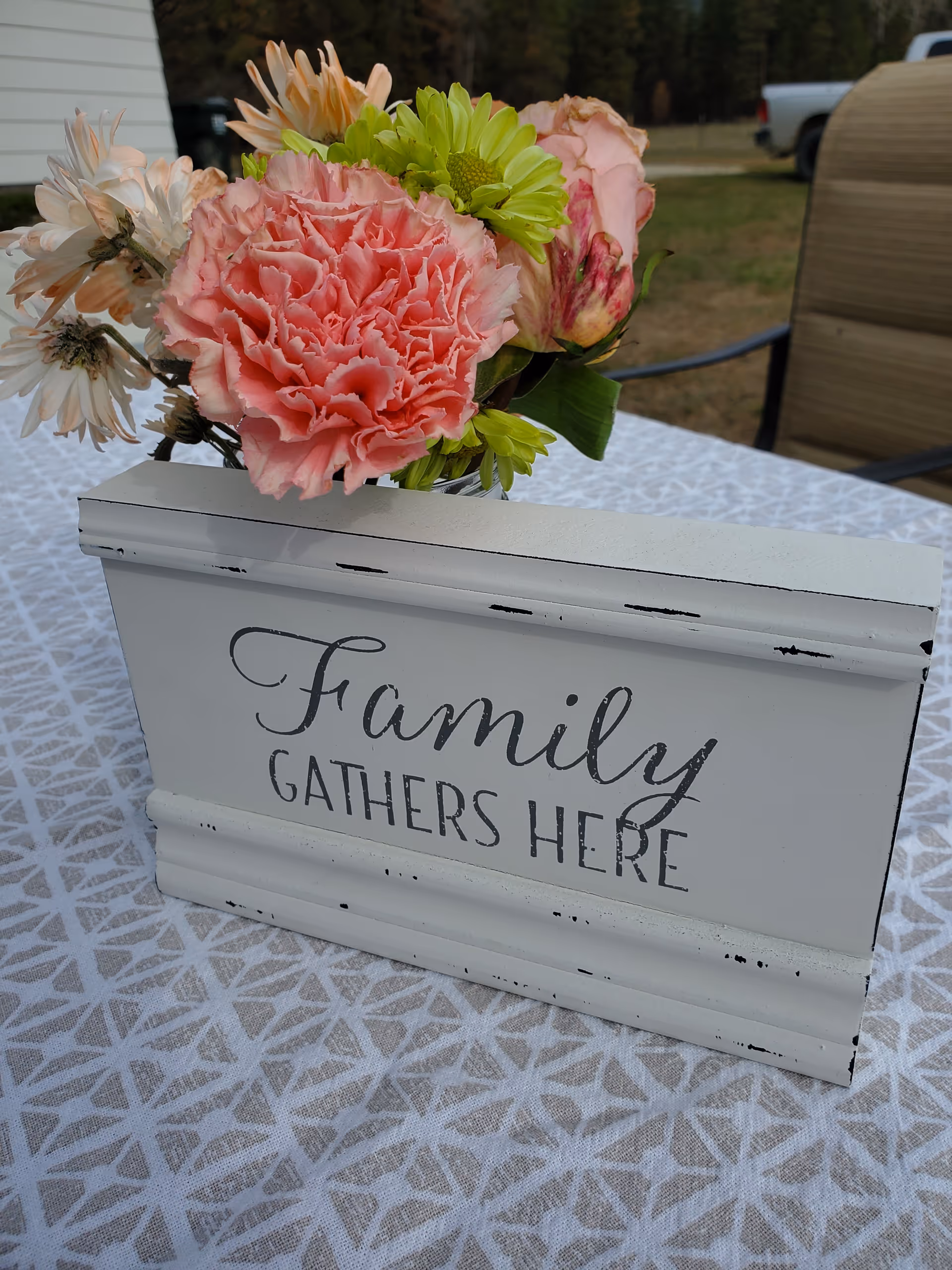 A white decorative wooden box with the words 'Family Gathers Here' written on it, holding a bouquet of flowers including pink carnations, green chrysanthemums, and other mixed flowers, placed on a patterned tablecloth outdoors with a chair and grassy area in the background.