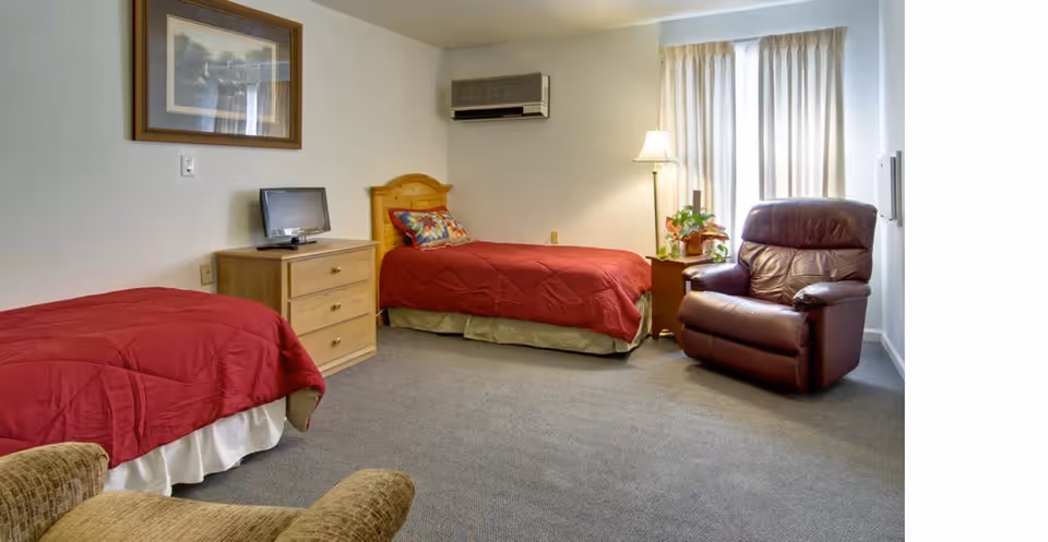 A senior living facility bedroom with two single beds covered in red bedspreads, a wooden dresser with a small TV on top, a brown leather recliner chair, a side table with a lamp and a plant, and a window with white curtains.