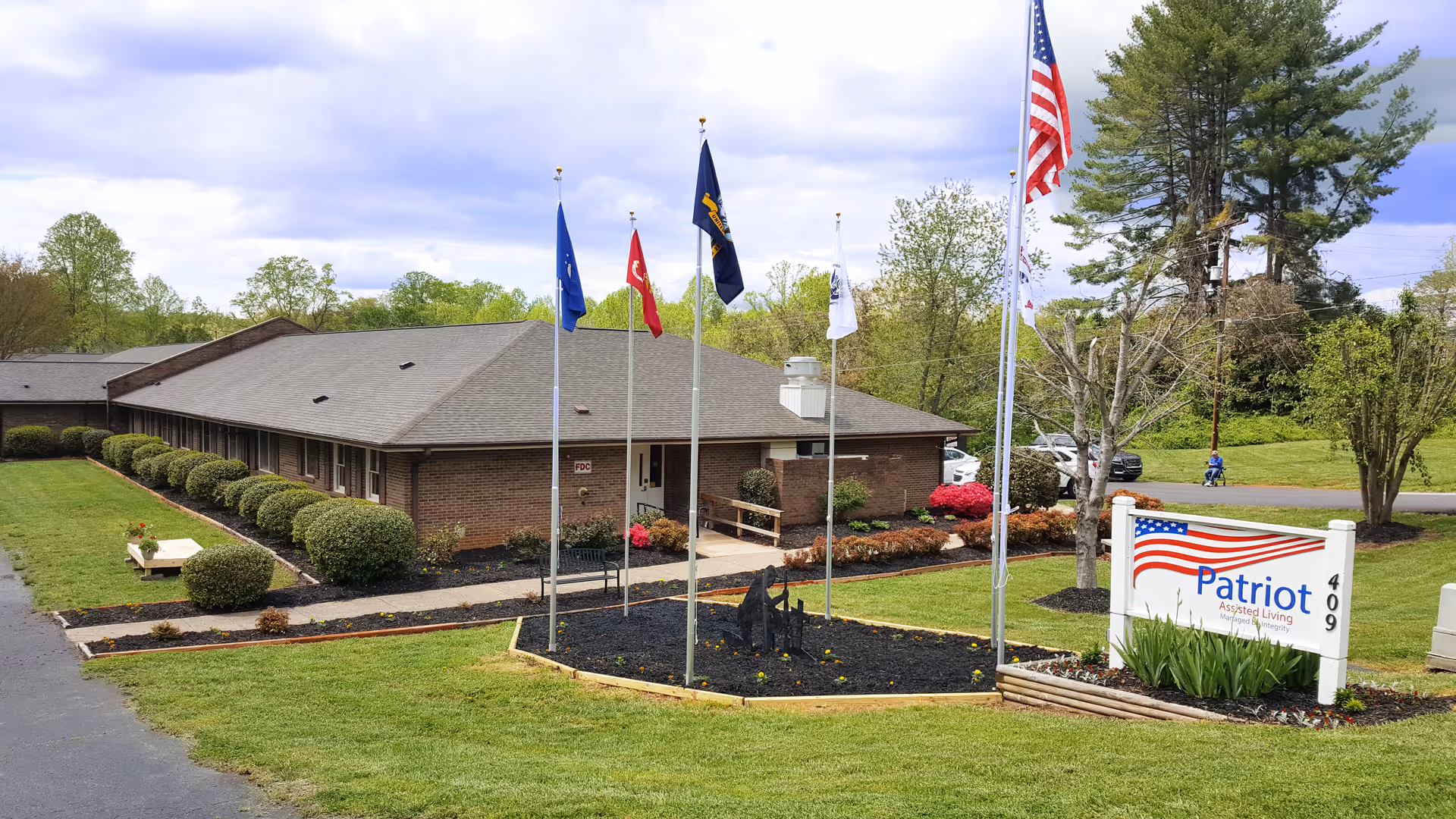 Exterior view of Patriot Assisted Living facility showing a single-story brick building with a gray roof, surrounded by well-maintained green lawns, bushes, and trees. There are five flagpoles with various flags, including the American flag, in front of the building near a sign that reads 'Patriot Assisted Living 409'.