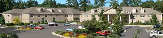 Exterior view of a large two-story assisted living facility building with multiple windows, surrounded by trees and landscaped greenery, with a parking lot in front containing several parked cars.