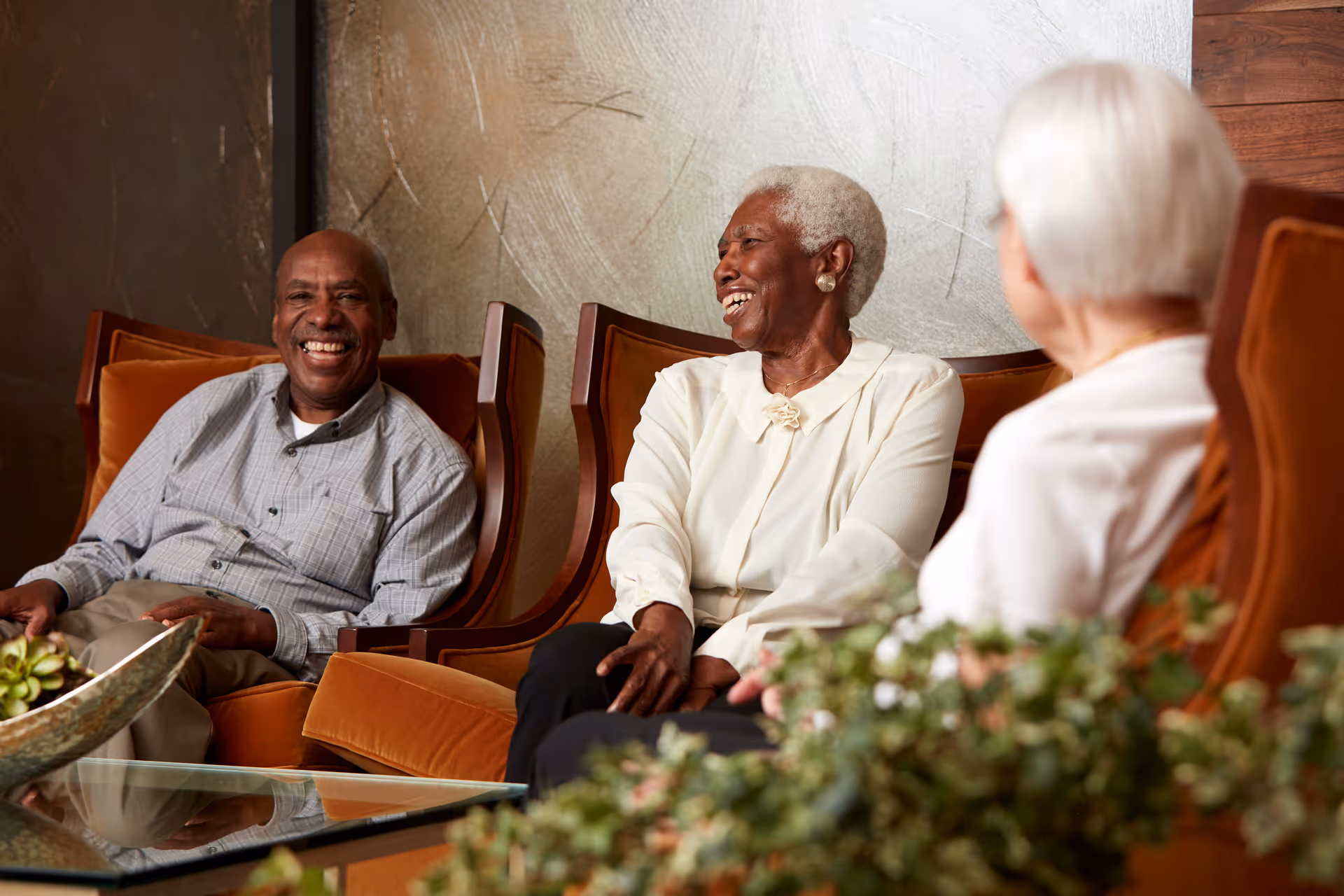 Three older adults sitting in a cozy lounge on orange armchairs, smiling and conversing.