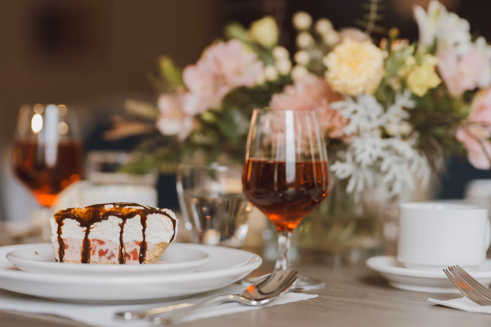 A close-up of a dessert plate with a slice of pie topped with whipped cream and chocolate drizzle, accompanied by a glass of red beverage, a white coffee cup, and a floral centerpiece in the background on a dining table.