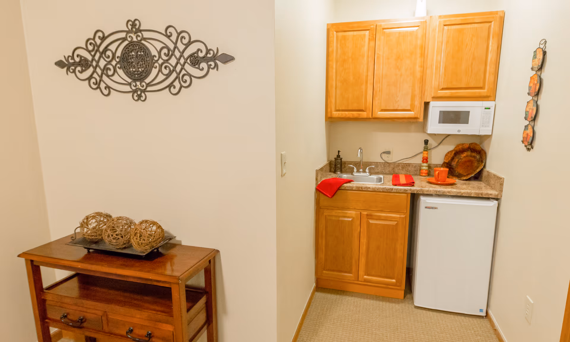 Small kitchenette with wooden cabinets, sink, microwave, mini-fridge, and a decorative wooden table with woven balls.