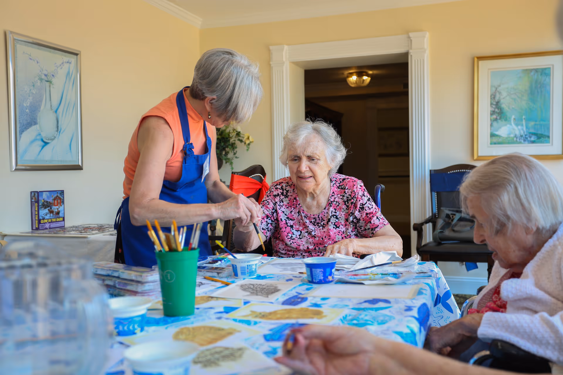 An elderly woman in a pink floral shirt is seated at a table covered with art supplies and papers, while a caregiver wearing a blue apron assists her with painting. Another elderly woman is seated nearby, also engaged in the activity. The room has light yellow walls with framed artwork and a doorway leading to another room.