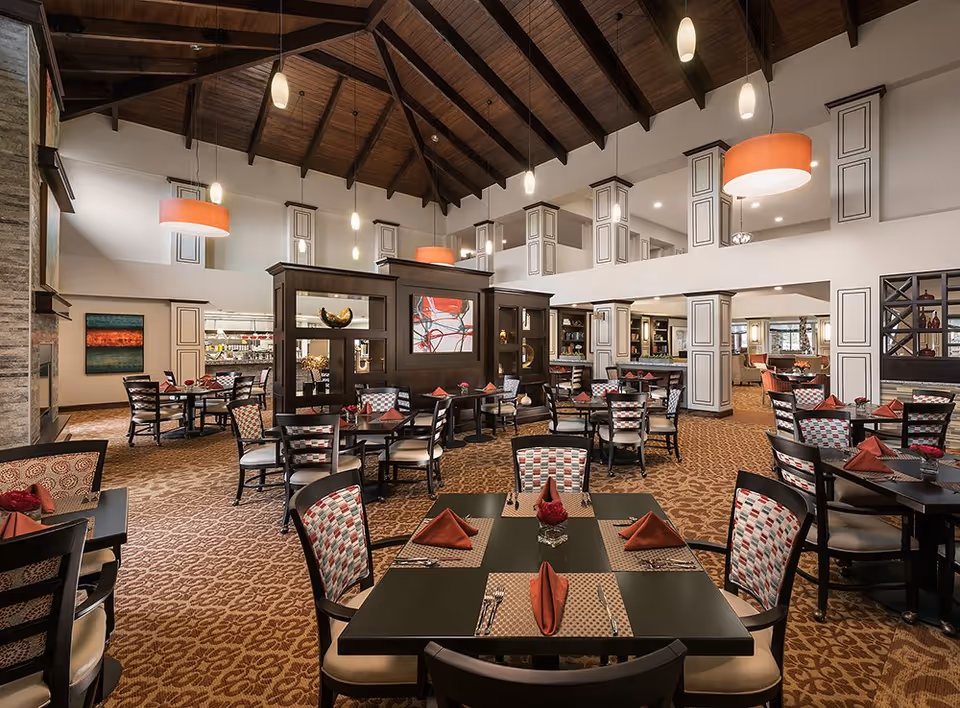 Spacious upscale dining room with multiple set tables, patterned chairs and carpeting, high wood-beamed ceiling, and pendant lighting.