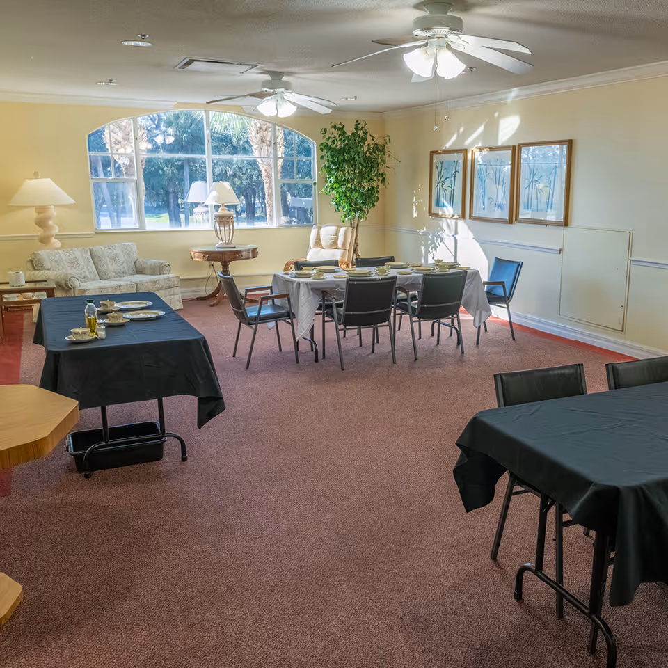 A well-lit dining room with several tables covered in black and white tablecloths, set with plates, cups, and utensils. There is a large window with an arched top letting in natural light, a beige sofa, a round wooden side table with a lamp, a potted plant, and three framed artworks on the wall. The room has ceiling fans with lights and carpeted flooring.