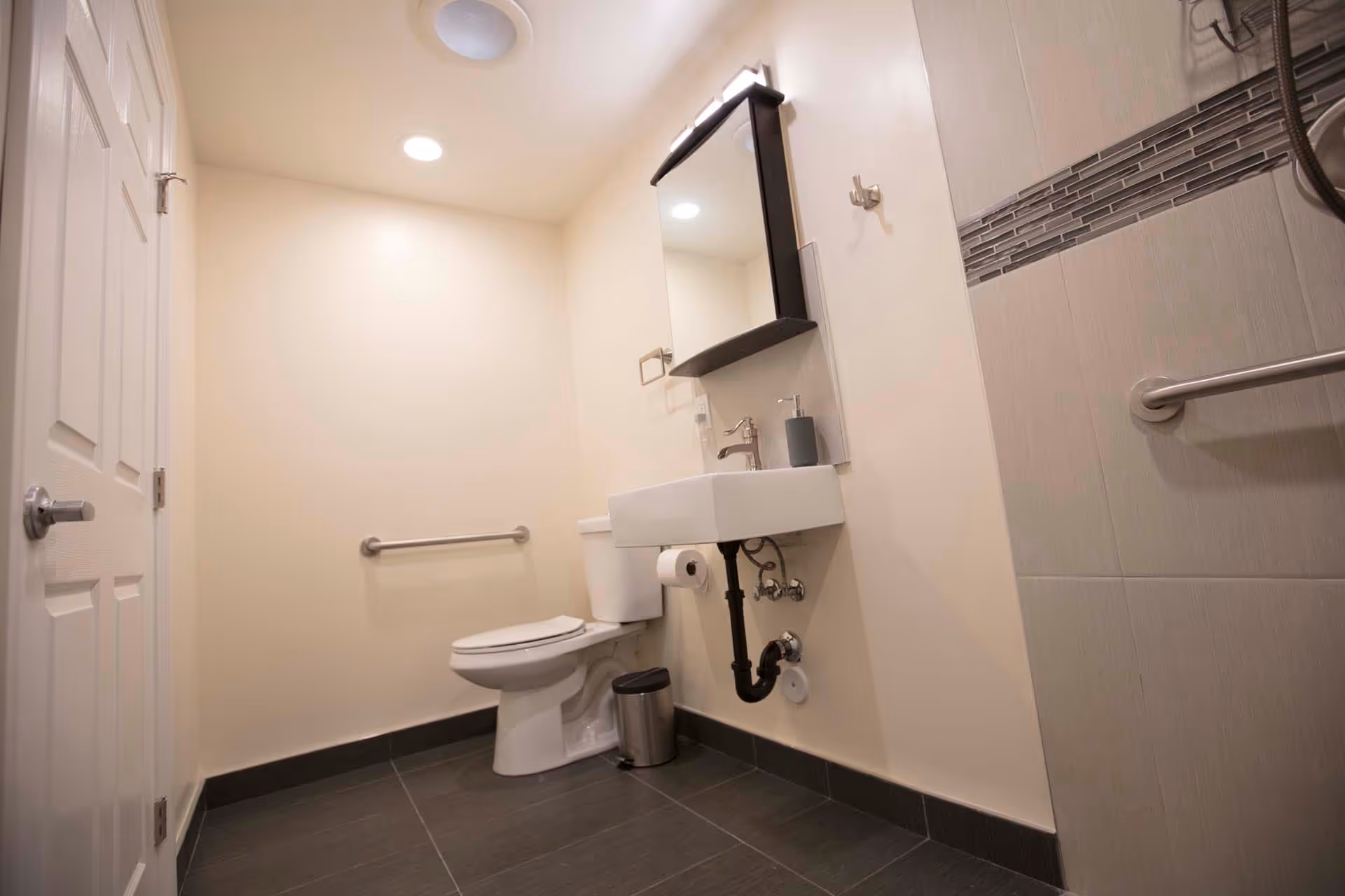 Accessible bathroom with a toilet, wall-mounted sink and mirror, grab bars, and tiled floor.