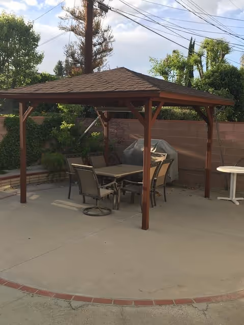 Outdoor patio area with a wooden gazebo covering a table and six chairs. There is a barbecue grill covered with a protective cover against a brick wall, surrounded by greenery and trees under a partly cloudy sky.