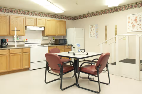 A kitchen area with wooden cabinets, a white stove, refrigerator, microwave, and coffee maker. In the center, there is a square table with four maroon cushioned chairs. The walls have a floral border near the ceiling and some anatomical charts. There is also a small white ramp with handrails on the right side.