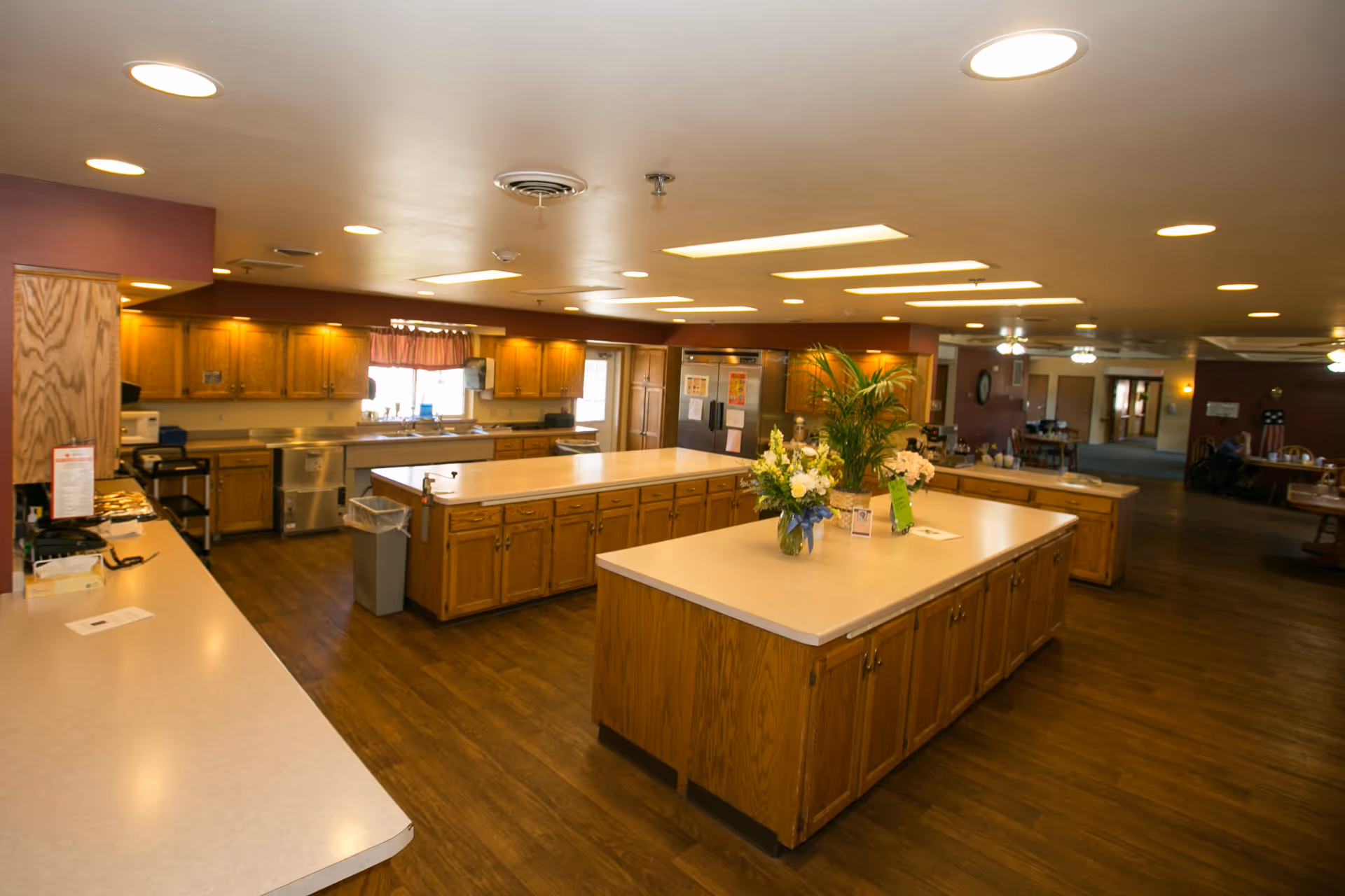 Spacious senior living facility kitchen with wooden cabinets, two large central islands with light-colored countertops, flowers and plants on one island, stainless steel appliances, and warm lighting.
