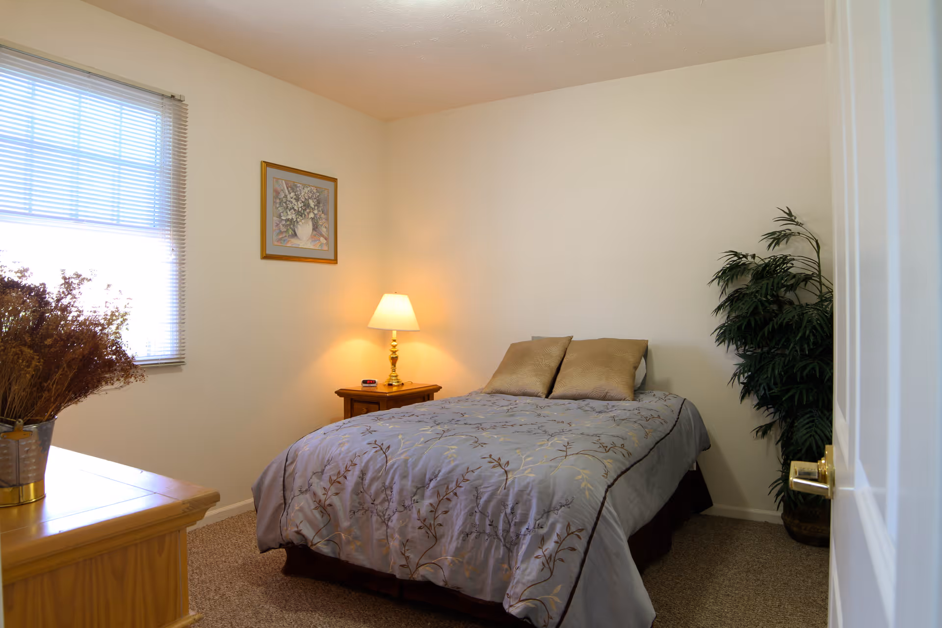 A cozy bedroom with a bed covered in a light blue and brown floral comforter, two beige pillows, a wooden nightstand with a lamp, a framed floral painting on the wall, a window with blinds, a wooden dresser with a vase of dried flowers, and a large green potted plant in the corner.