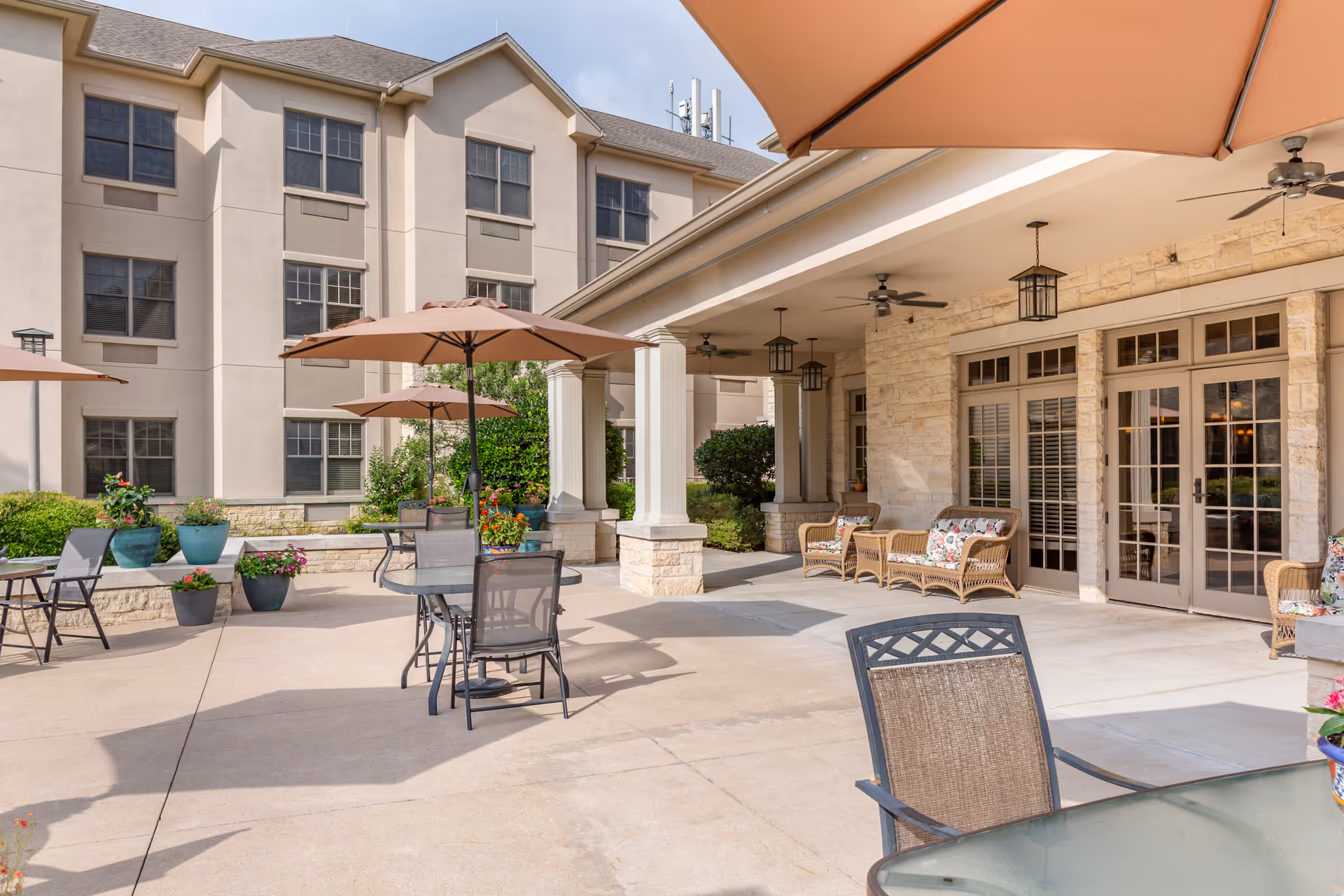 Outdoor patio area at Brookdale Northwest Hills featuring tables with umbrellas, chairs, potted plants, and a covered seating area with wicker furniture and ceiling fans attached to the building.