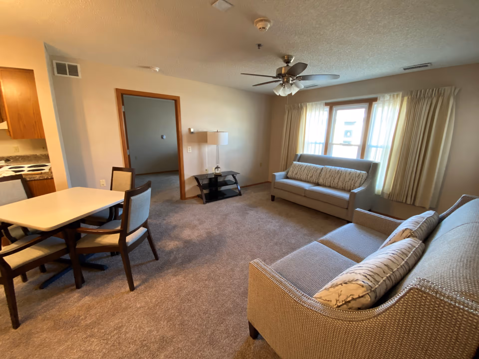 A living room area in a retirement center featuring two sofas with patterned cushions, a window with curtains allowing natural light, a ceiling fan with lights, a small black glass table with a lamp, and a dining table with four chairs adjacent to a kitchen area.
